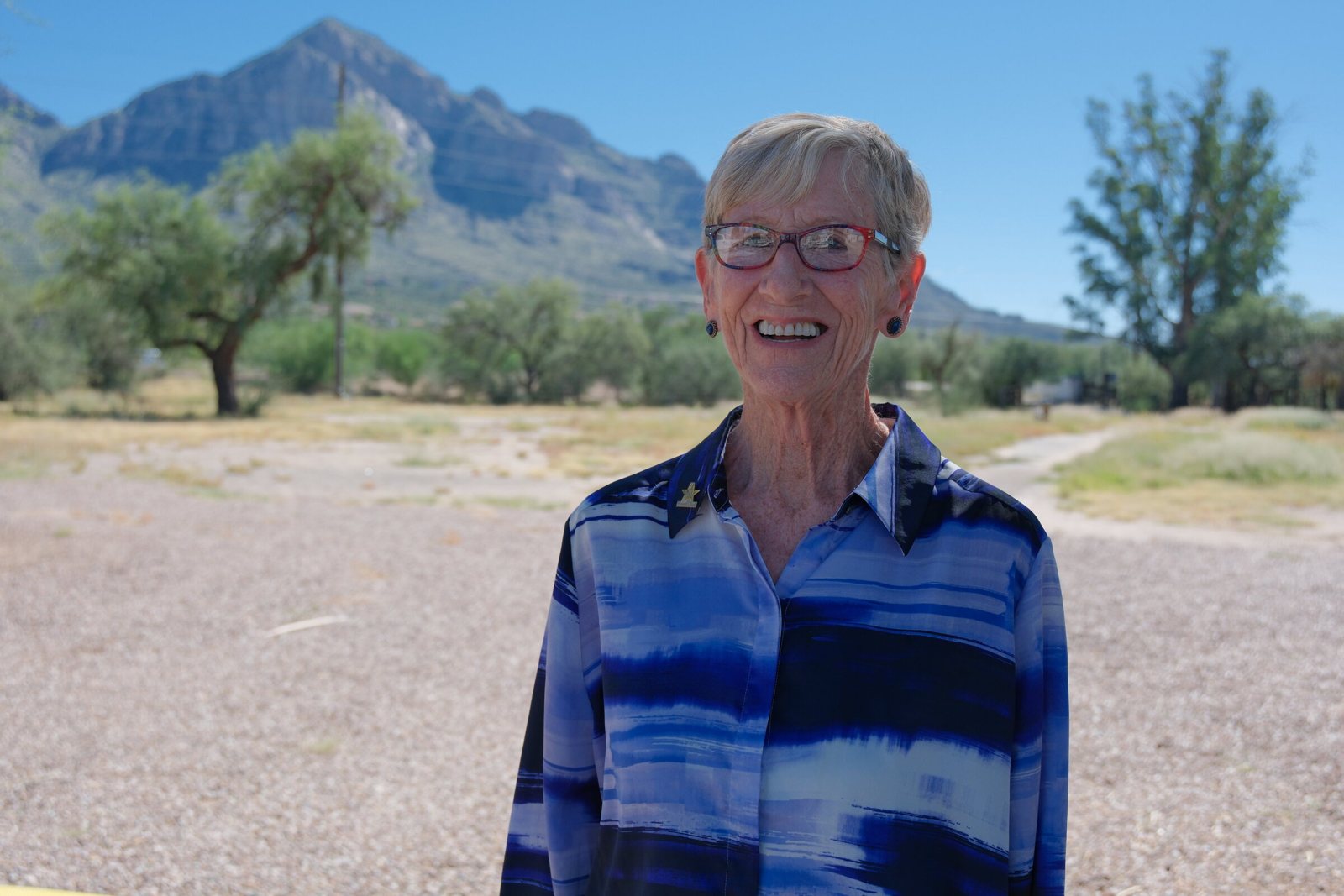 Devon Sloan, smiling, in a blue-and-white striped blouse, standing on a gravel lot with mesquite trees and Pusch Ridge behind her.